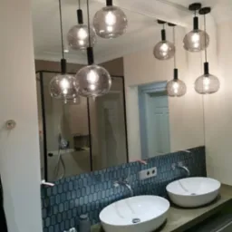 Nowoczesna łazienka z owalnymi umywalkami, mozaiką i dużym lustrem, w którym odbijają się szklane lampy wiszące.