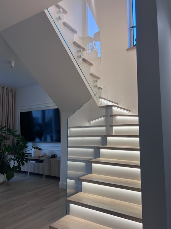 Nowoczesne schody z podświetleniem LED stopni i szklaną balustradą mocowaną punktowo, widok z dołu, w tle salon z telewizorem.