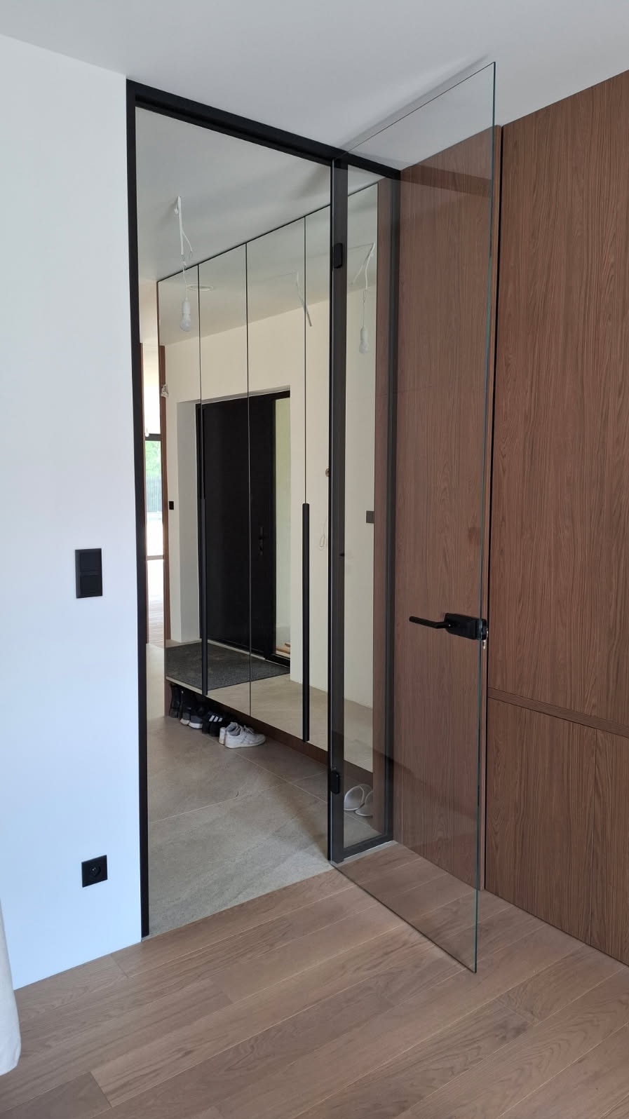 Nowoczesne drzwi szklane z czarną ramą i minimalistyczną klamką, oddzielające przestrzeń z lustrzaną szafą wnękową. Jasne wnętrze z drewnianą podłogą.