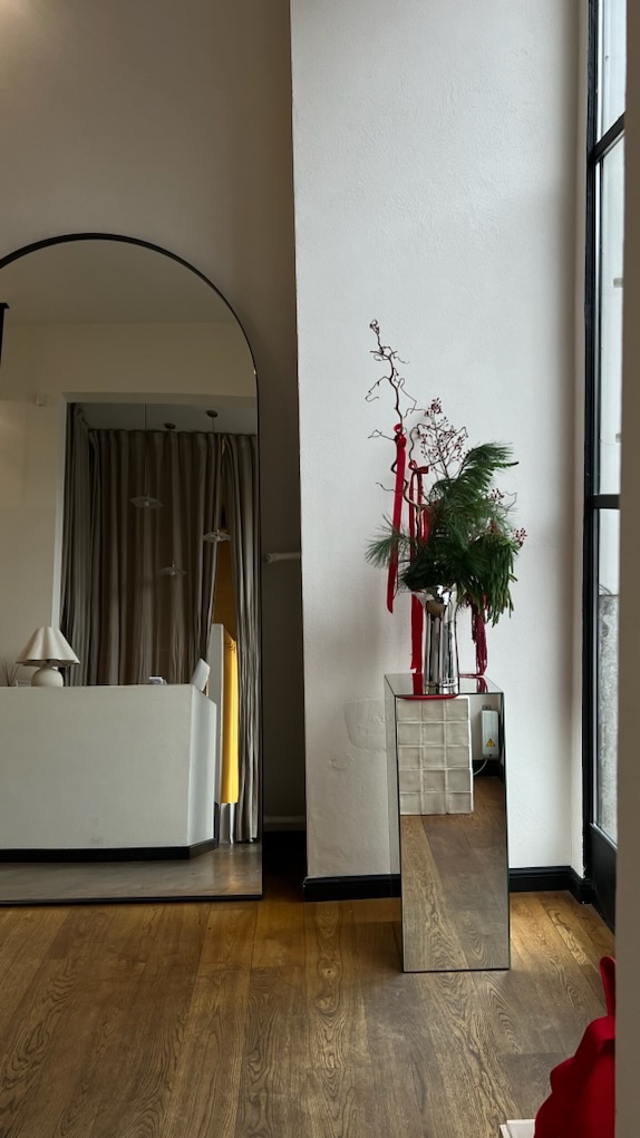 Eleganckie lustro łukowe w czarnej ramie odbija wnętrze salonu. Dekoracja świąteczna na lustrzanym postumencie dodaje uroku. Parkiet w ciepłym odcieniu brązu.