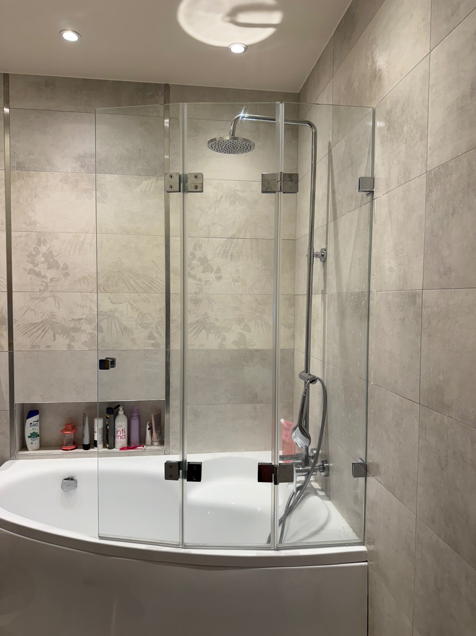 Nowoczesna kabina nawannowa ze szkła hartowanego, z chromowanymi zawiasami, doświetlona halogenami, w łazience z beżowymi płytkami z motywem roślinnym.