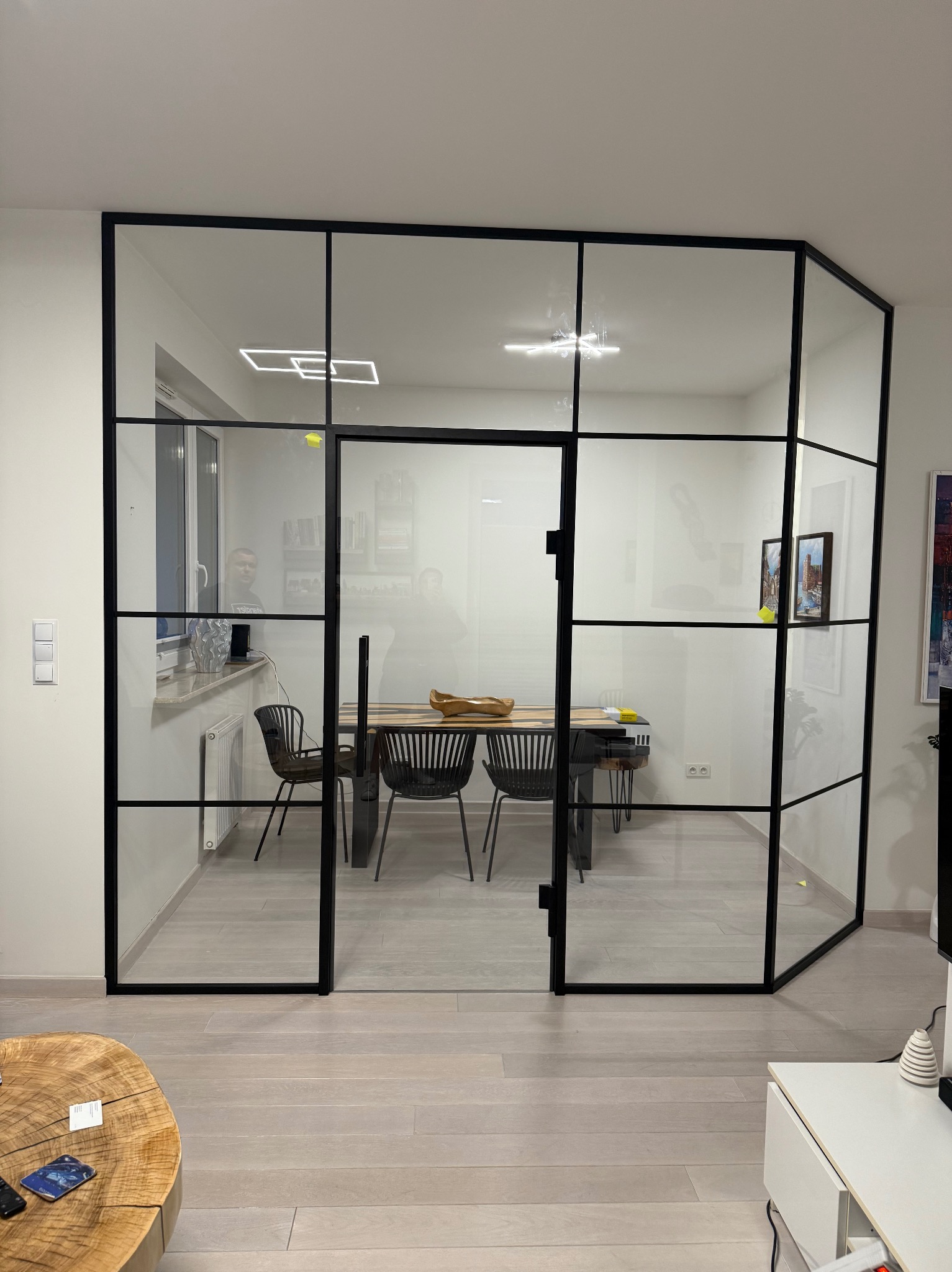 Nowoczesne drzwi wewnętrzne szklane z czarnymi szprosami oddzielają salon od jadalni. Widoczny stół i krzesła w jadalni, minimalistyczny wystrój.