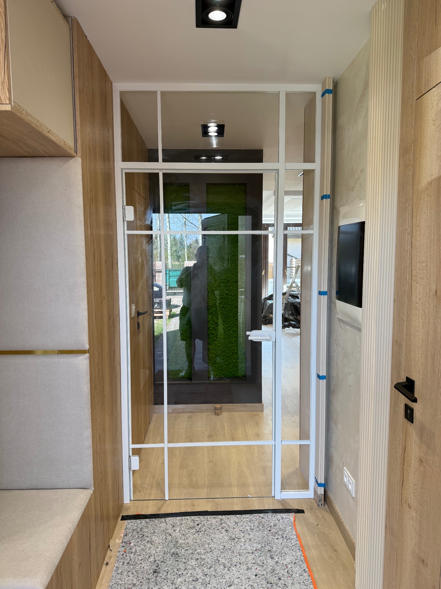 Nowoczesne drzwi z białym szkieletem i przezroczystym szkłem, z widokiem na zieloną ścianę w tle. Minimalistyczny design w jasnym wnętrzu.