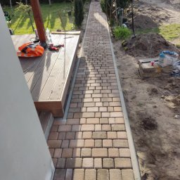 Brukersi - Ułożona ścieżka z kostki brukowej w odcieniach beżu i brązu prowadzi od drewnianego tarasu, obok którego leży pomarańczowa kamizelka odblaskowa, wzdłuż rabaty z tujami; widoczny młotek brukarski.