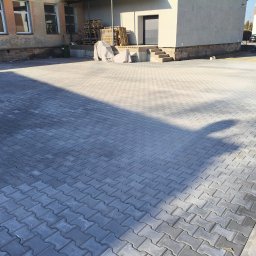 Brukersi - Szara kostka brukowa ułożona na dużej powierzchni przed budynkiem. Widoczne palety pod plandeką, schody i zaparkowany samochód w tle. Słoneczny dzień.