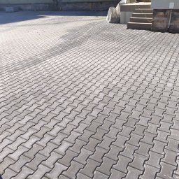 Brukersi Arkadiusz Joński - Ułożona szara kostka brukowa typu 'I' na podjeździe, widok z góry, z fragmentem schodów i budynku w tle. Regularny wzór, równa powierzchnia, świeżo po realizacji.