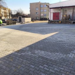 Brukersi - Szara kostka brukowa ułożona na placu, z widocznym budynkiem w tle i osobą zamiatającą. Część placu oświetlona słońcem, tworząca kontrast z zacienioną strefą.