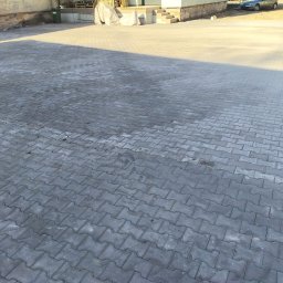 Brukersi Arkadiusz Joński - Ułożona szara kostka brukowa typu 'I', tworząca dużą powierzchnię placu przed budynkiem. Widoczne nierównomierne oświetlenie i cień. W tle budynek i zaparkowany samochód.