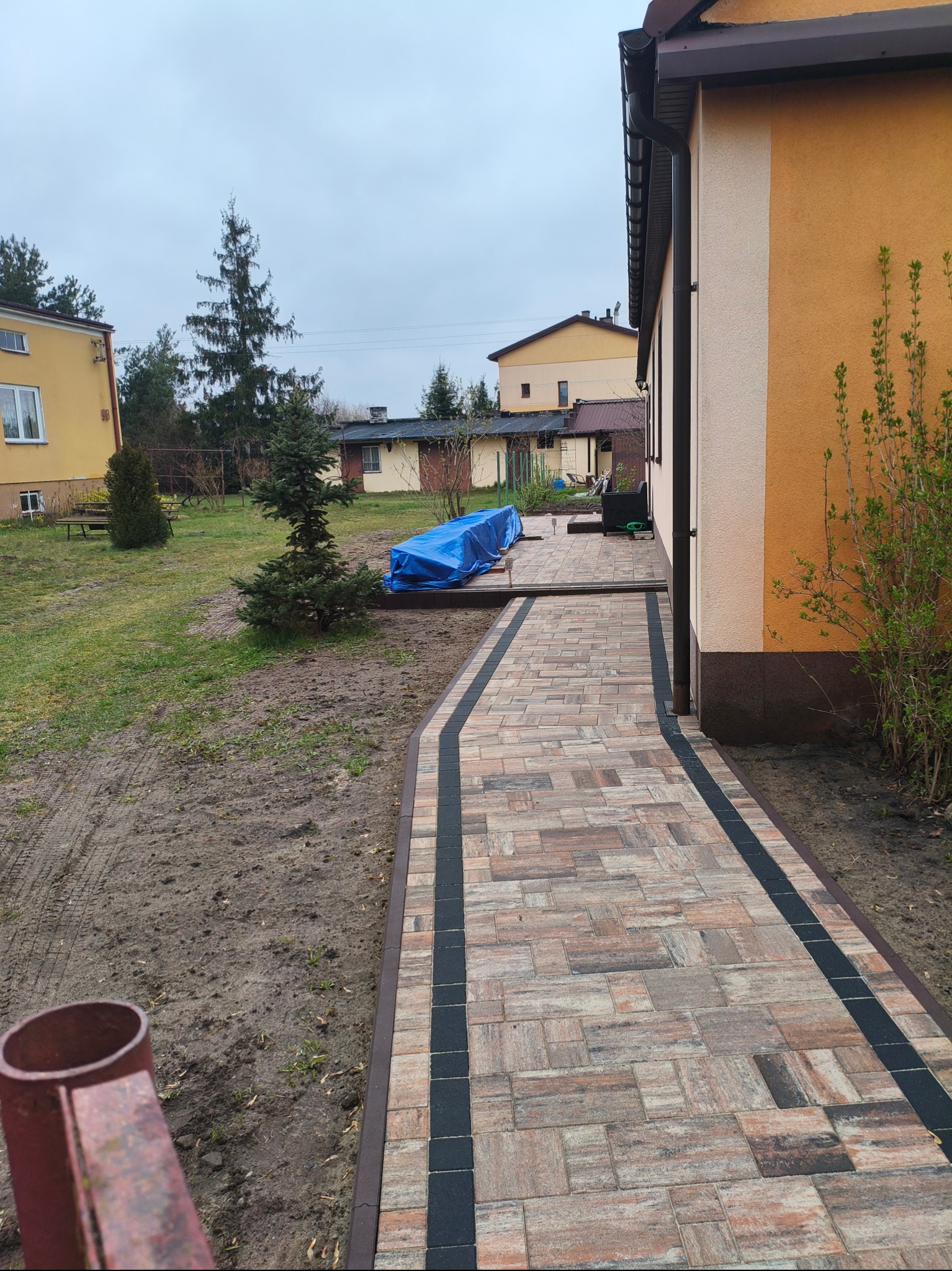 Ścieżka z kostki brukowej w odcieniach brązu i czerni prowadząca wzdłuż ściany budynku, na trawniku widoczna niebieska plandeka przykrywająca materiały budowlane.