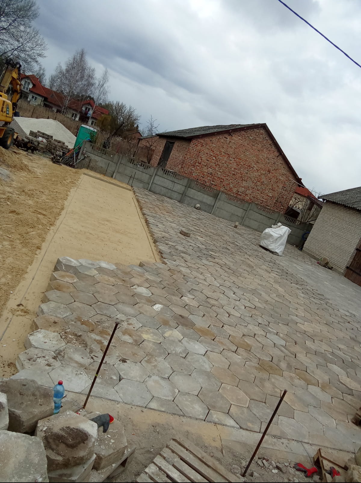 Układanie heksagonalnej kostki brukowej na posesji, widoczny fragment podsypki piaskowej, paleta kostki, butelka wody i narzędzia pracy, w tle budynki mieszkalne i pochmurne niebo.