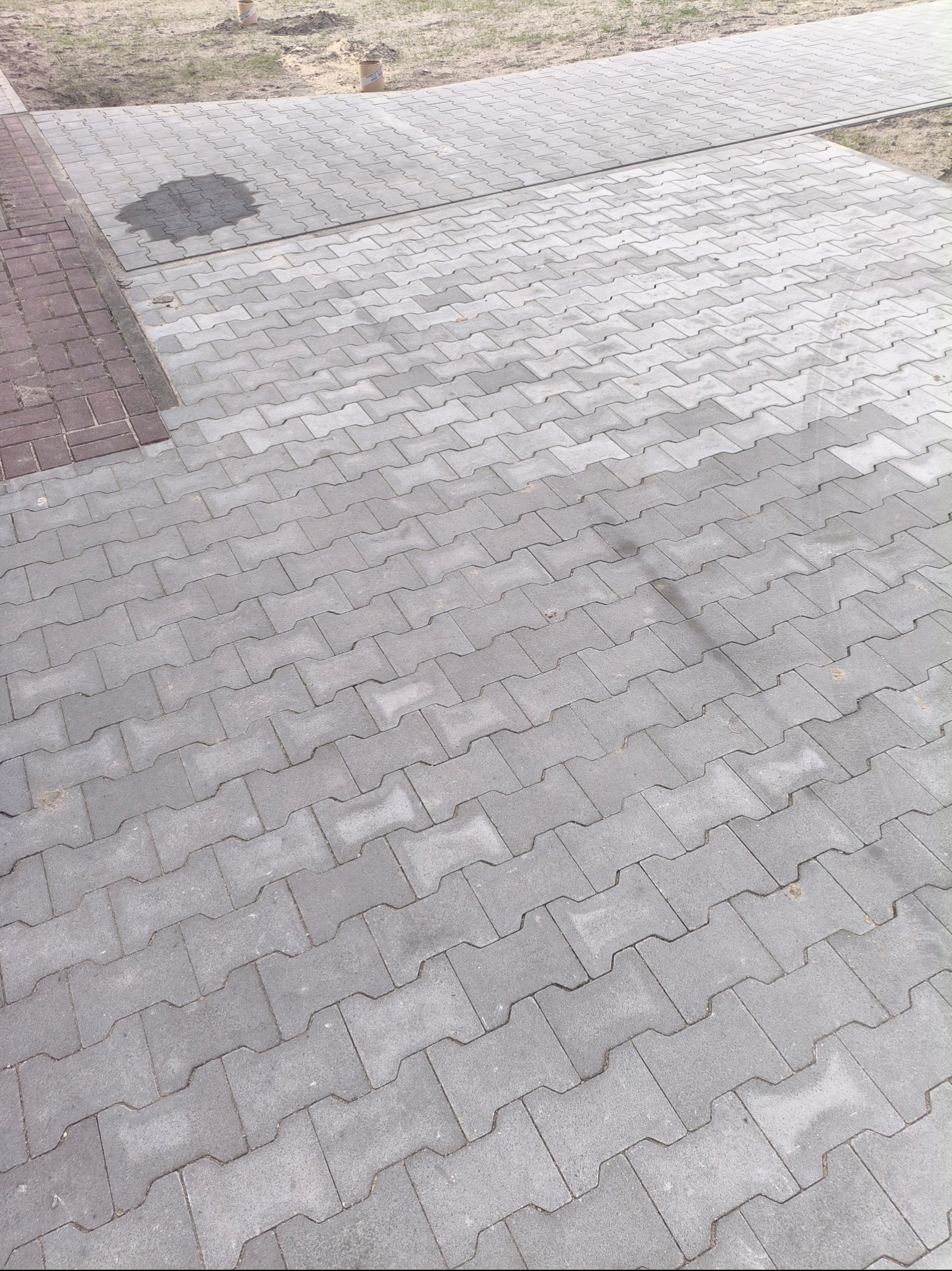 Szara kostka brukowa ułożona na podjeździe, widoczna plama po deszczu i fragment czerwonej kostki po lewej stronie, w tle ziemia i dwa kartonowe cylindry.