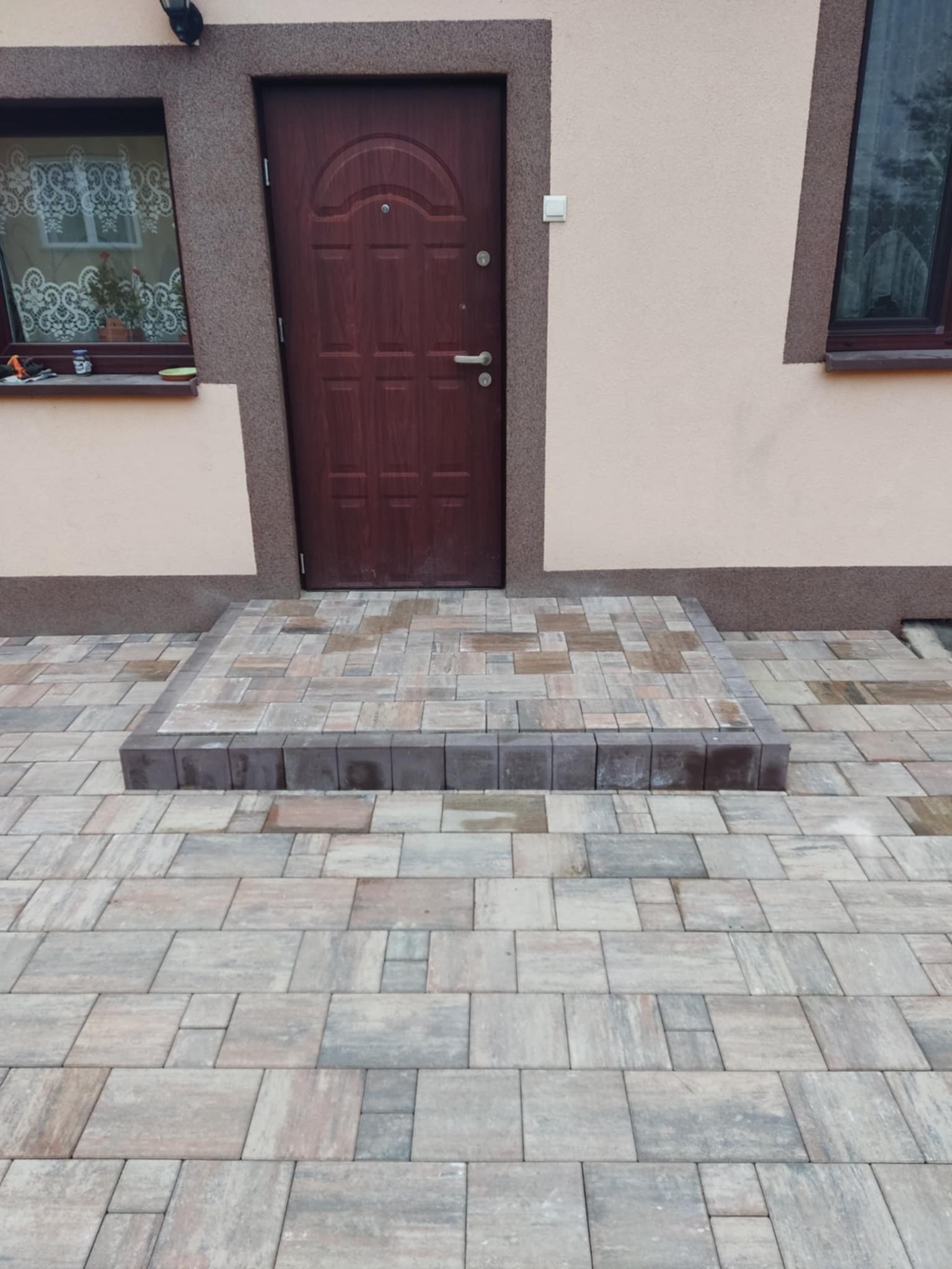 Ułożona kostka brukowa prowadząca do drzwi wejściowych domu, widoczne schody wykończone ciemniejszą kostką.