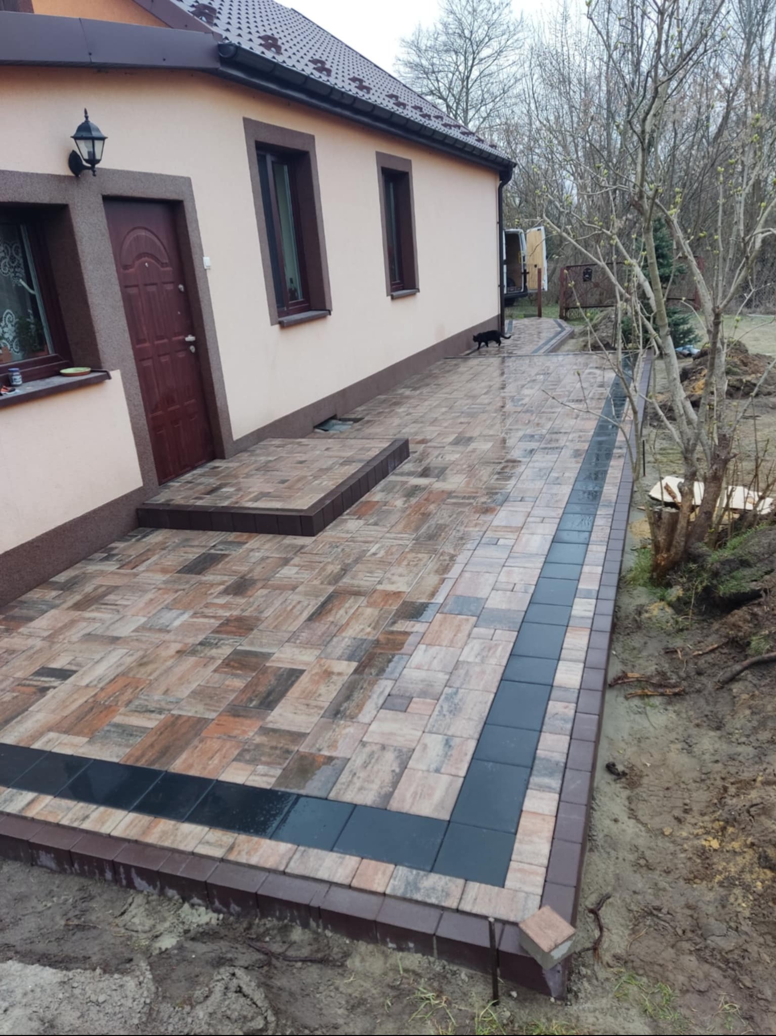 Ułożona kostka brukowa w odcieniach brązu i beżu z ciemnoszarym pasem biegnącym wzdłuż ścieżki przed domem, widoczny czarny kot spacerujący po kostce, budynek z beżową elewacją i brązowymi...