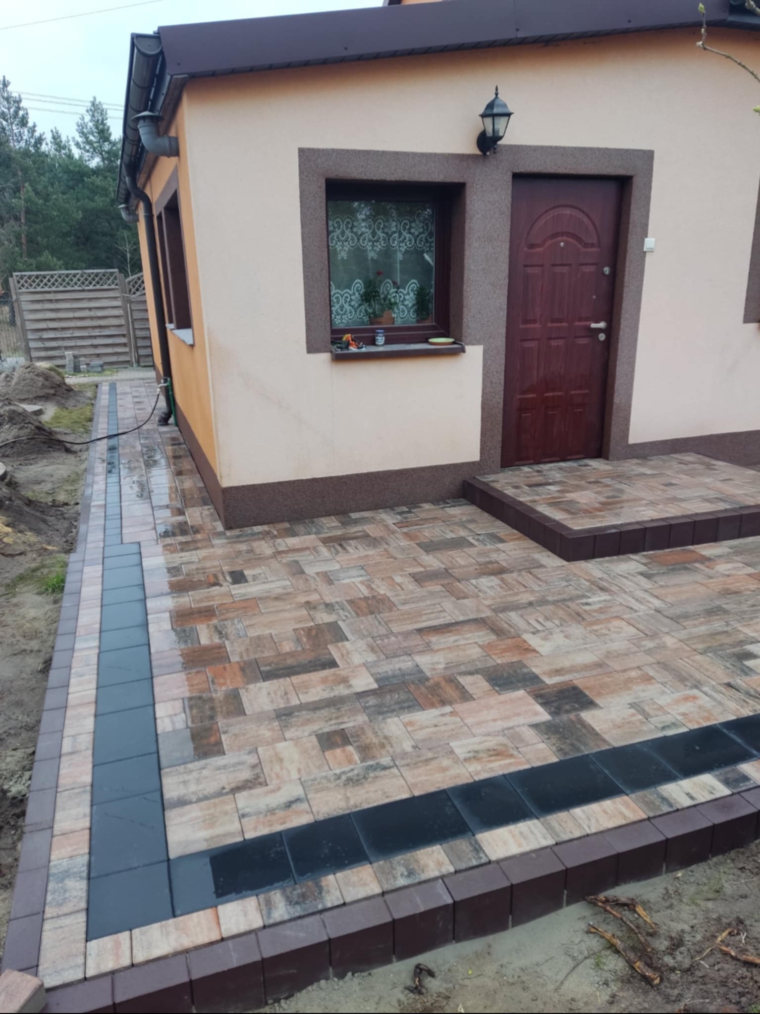 Ułożona kostka brukowa w odcieniach brązu i beżu tworząca ścieżkę i podest przed domem, obrzeża z ciemnej kostki, widoczna elewacja budynku w kolorze beżowym.