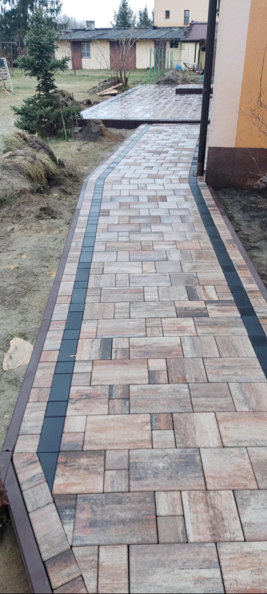Ułożona ścieżka z kostki brukowej o nieregularnym kształcie i kolorach ziemi, obramowana ciemnymi kostkami i brązowym obrzeżem, prowadząca wzdłuż budynku z widocznym ogrodem w tle.