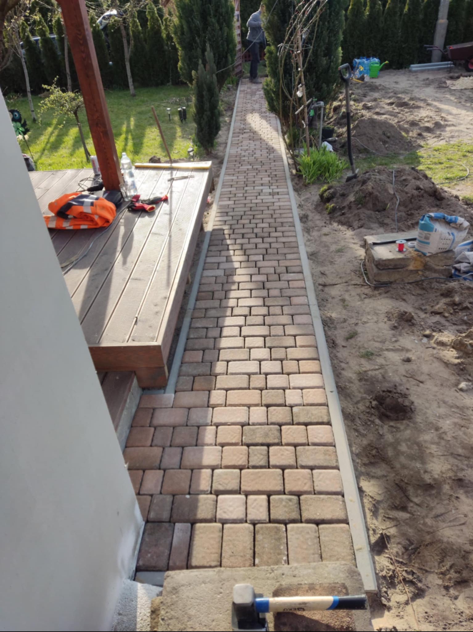 Ułożona ścieżka z kostki brukowej w odcieniach beżu i brązu prowadzi od drewnianego tarasu, obok którego leży pomarańczowa kamizelka odblaskowa, wzdłuż rabaty z tujami; widoczny młotek brukarski.