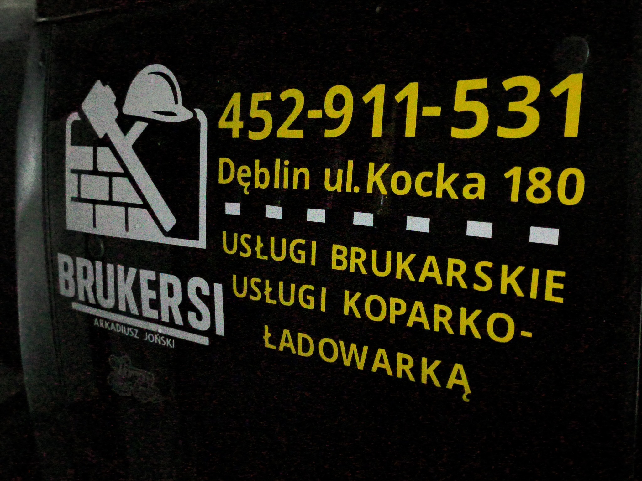 Reklama firmy brukarskiej z Dęblina: numer telefonu, adres, usługi brukarskie i koparko-ładowarką. Logo z kielnią, młotkiem i kaskiem na tle muru.