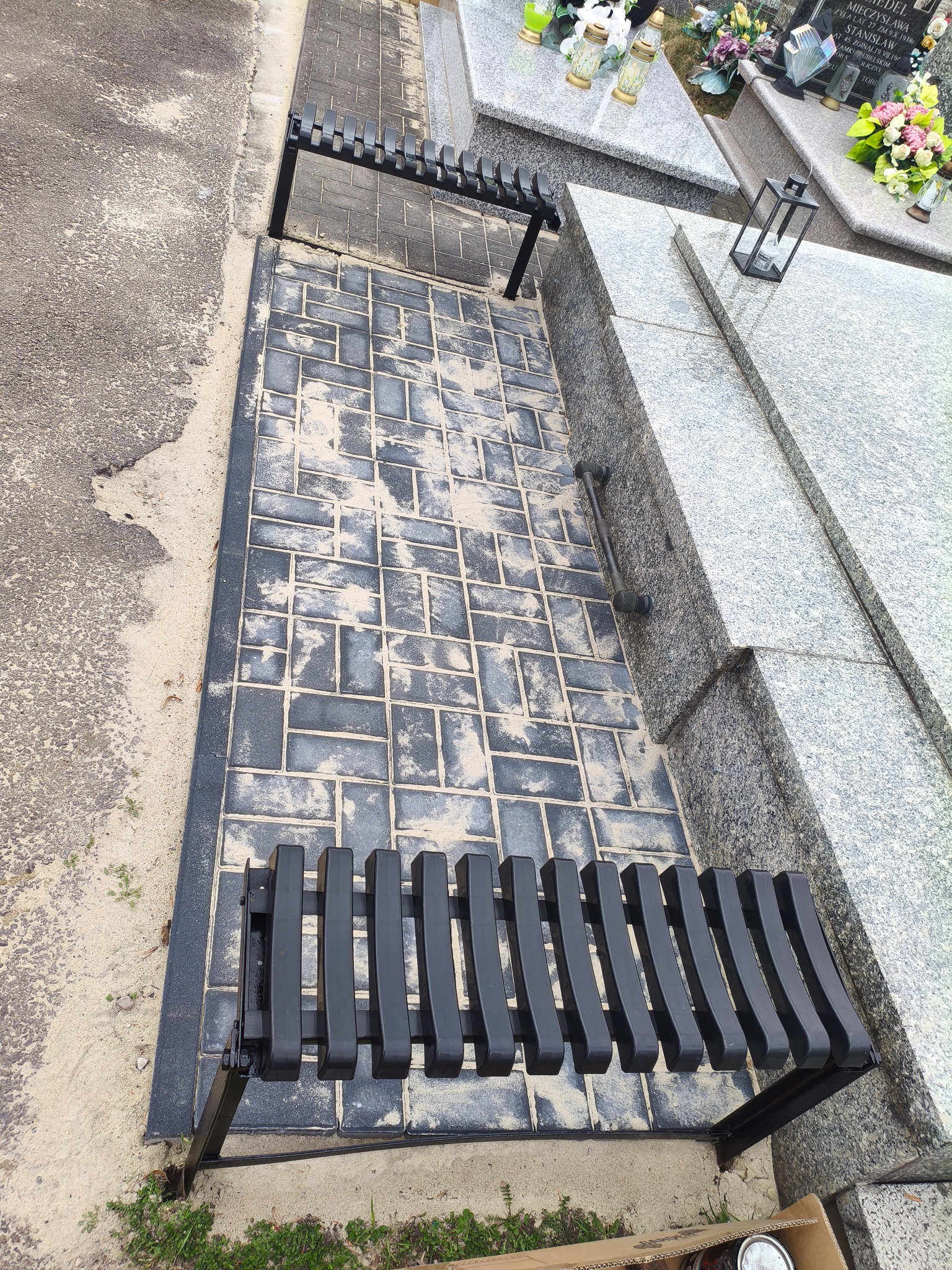 Ułożona kostka brukowa na cmentarzu, tworząca ścieżkę między grobami. Widoczna czarna ławka i fragmenty nagrobków z jasnego kamienia. Całość utrzymana w stonowanej kolorystyce.