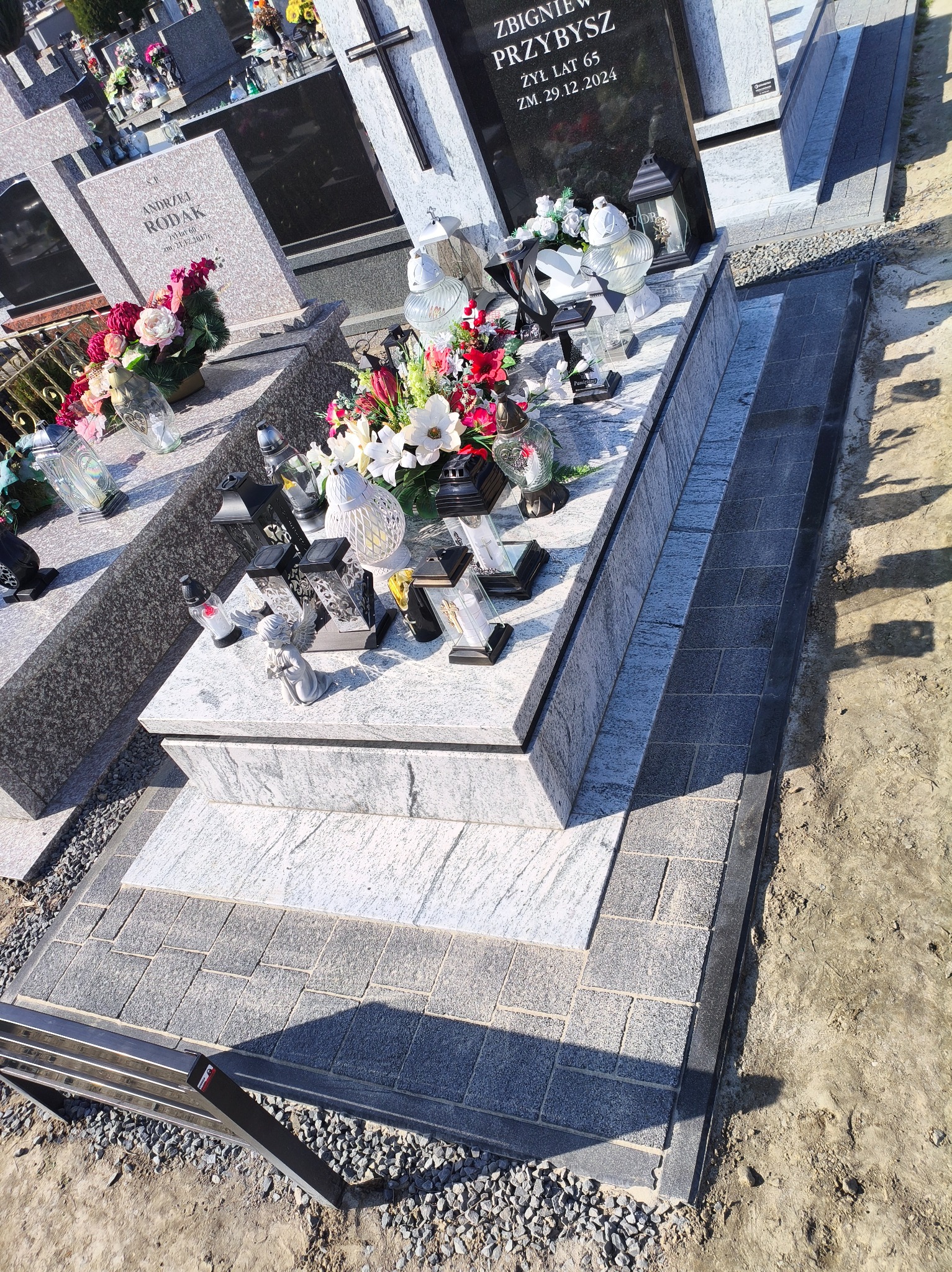 Granitowy nagrobek z kwiatami i lampionami, otoczony kostką brukową. Widok z góry, ukazujący detale kamieniarskie i kompozycję.