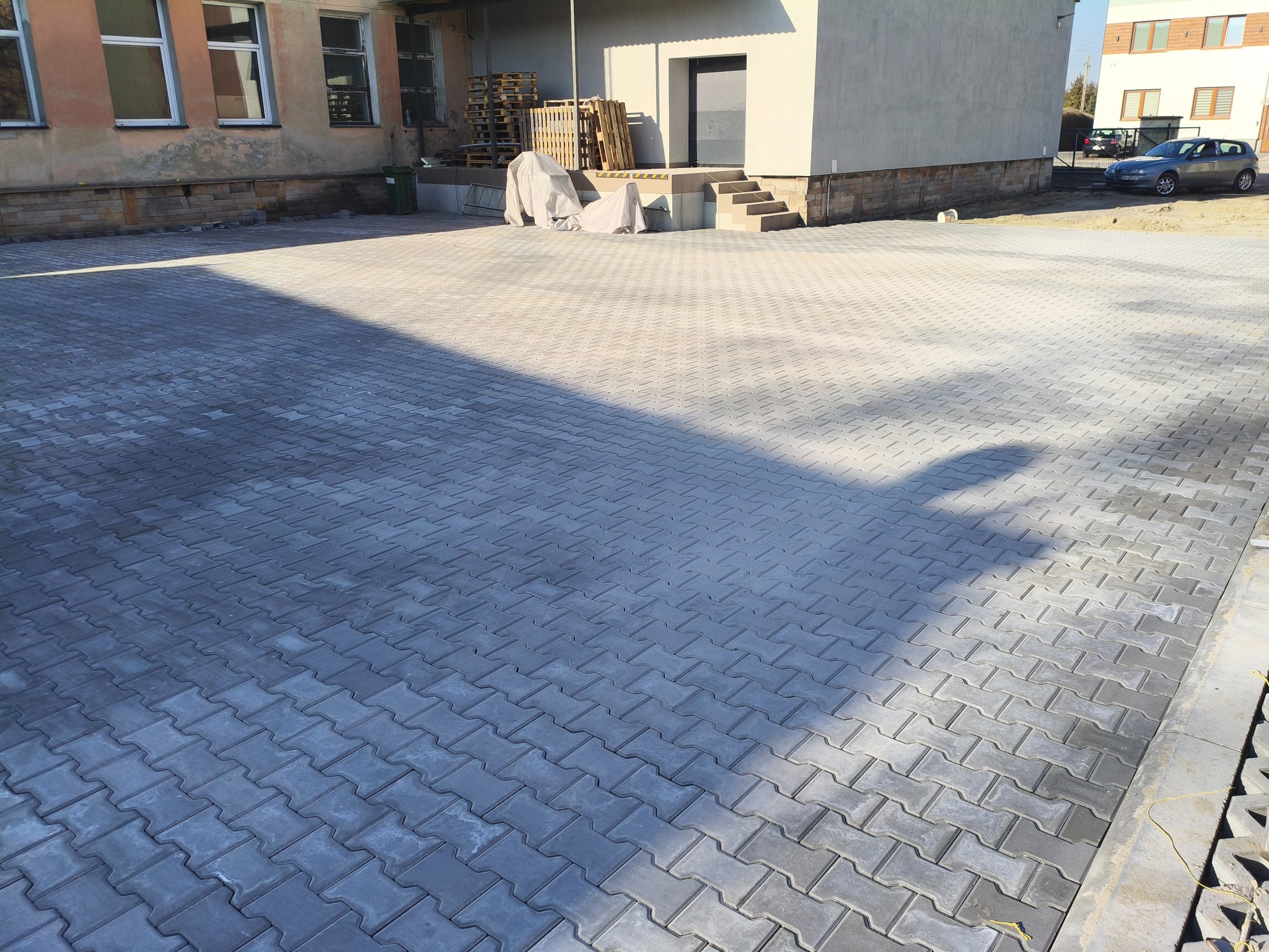 Szara kostka brukowa ułożona na dużej powierzchni przed budynkiem. Widoczne palety pod plandeką, schody i zaparkowany samochód w tle. Słoneczny dzień.