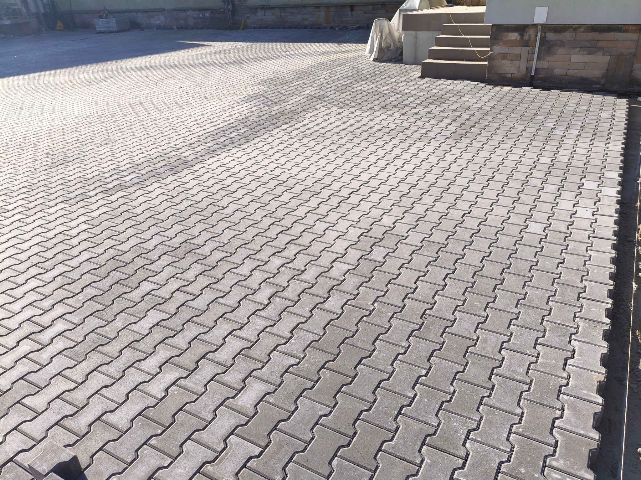 Ułożona szara kostka brukowa typu 'I' na podjeździe, widok z góry, z fragmentem schodów i budynku w tle. Regularny wzór, równa powierzchnia, świeżo po realizacji.