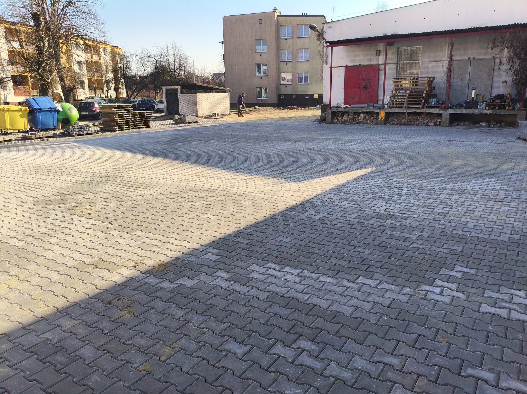 Szara kostka brukowa ułożona na placu, z widocznym budynkiem w tle i osobą zamiatającą. Część placu oświetlona słońcem, tworząca kontrast z zacienioną strefą.