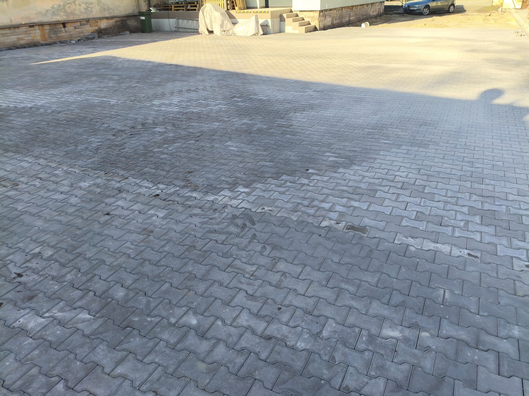 Ułożona szara kostka brukowa typu 'I', tworząca dużą powierzchnię placu przed budynkiem. Widoczne nierównomierne oświetlenie i cień. W tle budynek i zaparkowany samochód.