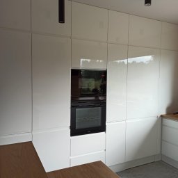 WAMA Wojciech Świątek - Biała, lakierowana zabudowa kuchenna z piekarnikiem Amica, oświetleniem punktowym i blatem z drewna w ciepłym odcieniu. Nowoczesny design z minimalistycznymi uchwytami.