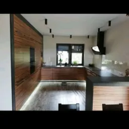 Nowoczesna kuchnia z drewnianymi frontami, czarnymi akcentami i podświetleniem LED pod szafkami. Widoczne okno z roślinami i czarny okap.