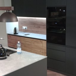 WAMA Wojciech Świątek - Nowoczesna kuchnia: czarne szafki, drewniane akcenty, marmurowy blat wyspy. Oświetlenie LED podkreśla unikalny design. Widoczny czajnik i butelka wody.
