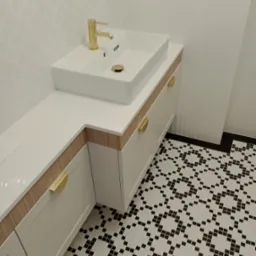 Biała szafka łazienkowa na wymiar z blatem, złotą armaturą i uchwytami, na tle wzorzystej podłogi. Umywalka prostokątna, nablatowa.