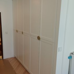 WAMA Wojciech Świątek - Elegancka, biała szafa wnękowa z subtelnymi, złotymi uchwytami, dopasowana do wnętrza z parkietem. Widoczne detale wykończenia i minimalistyczny design.