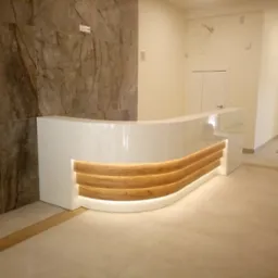 Błyszcząca, biała lada recepcyjna z elementami drewna i podświetleniem LED na tle marmurowej ściany. Widok na jasne wnętrze z drzwiami.