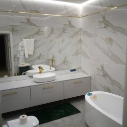 WAMA Wojciech Świątek - Elegancka łazienka z marmurowymi płytkami, białą szafką pod umywalkę z złotymi uchwytami i owalną wanną. Widoczne lustro i akcesoria łazienkowe.