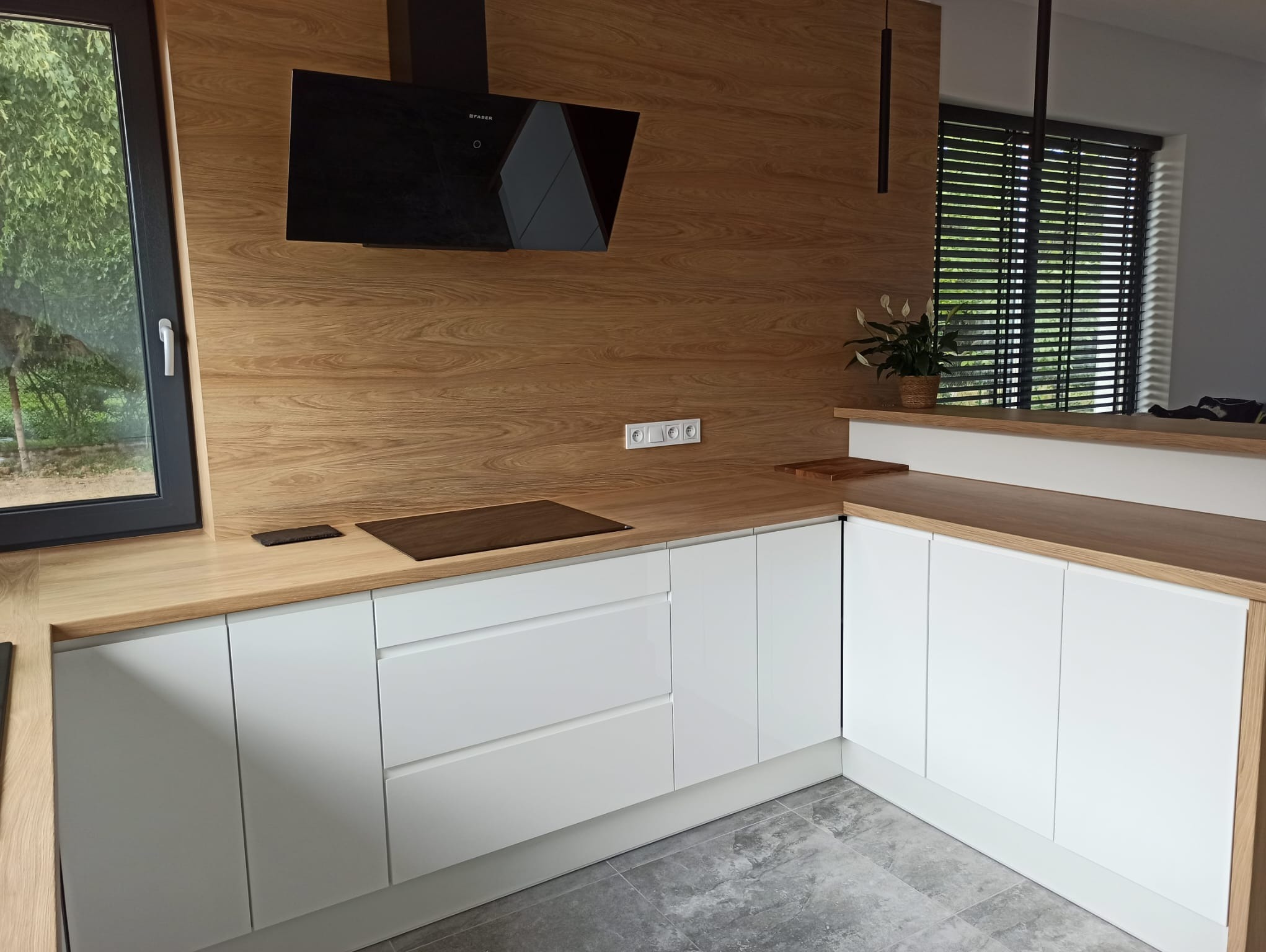 Nowoczesna kuchnia z białymi frontami i drewnianym blatem, czarny okap, widok na zewnątrz przez okno. Minimalistyczny design z elementami dekoracyjnymi.