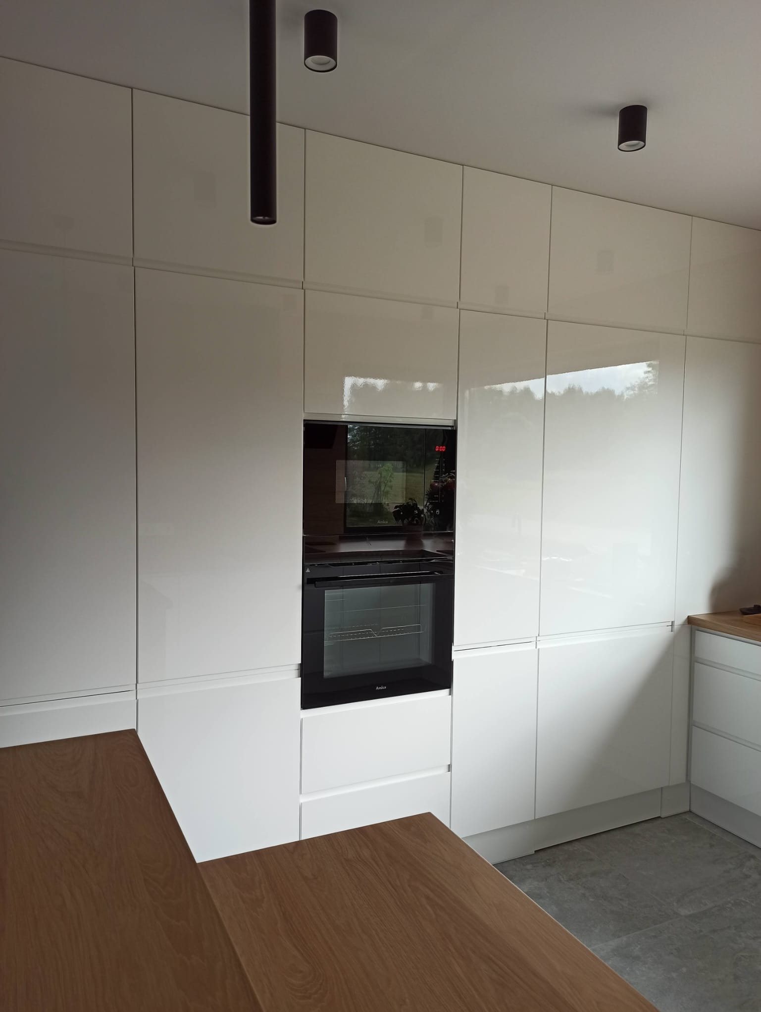 Biała, lakierowana zabudowa kuchenna z piekarnikiem Amica, oświetleniem punktowym i blatem z drewna w ciepłym odcieniu. Nowoczesny design z minimalistycznymi uchwytami.