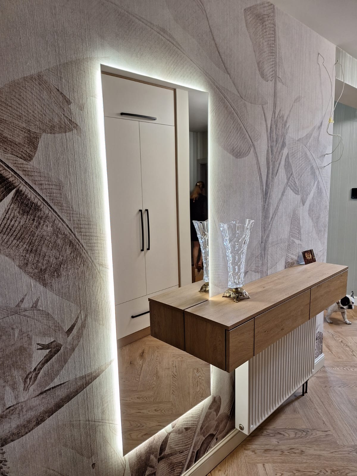 Nowoczesne wnętrze z szafą wnękową w lustrzanym odbiciu, podświetlanym LED, z drewnianą konsolą i wzorem liści na tapecie. Uwagę przykuwa pies w tle.