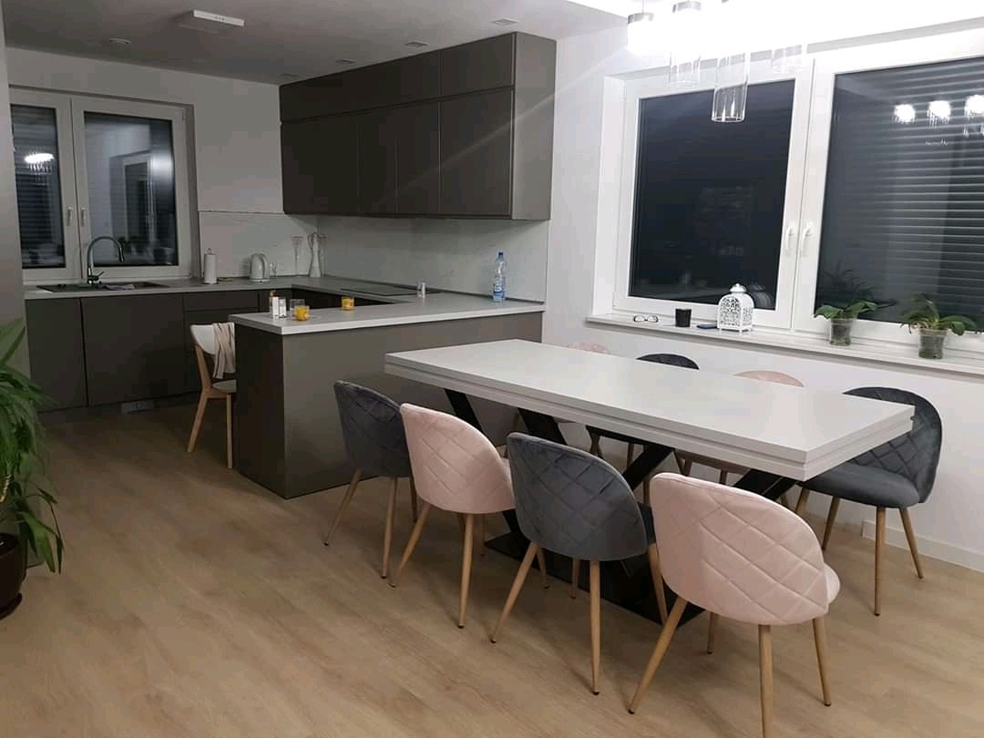 Nowoczesna kuchnia z ciemnymi szafkami i blatem, jasny stół z krzesłami w pastelowych kolorach. Minimalistyczny design, okno z roletami, drewniana podłoga.