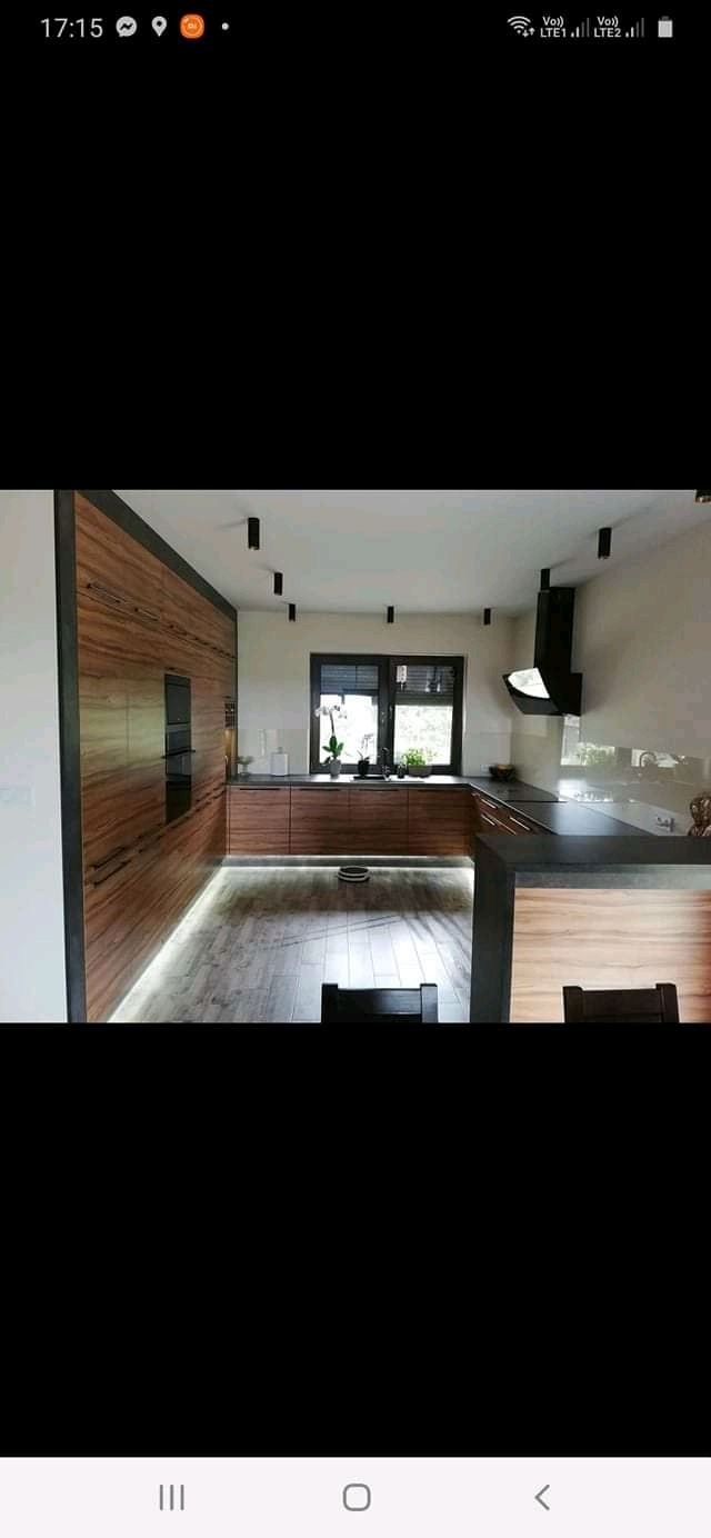 Nowoczesna kuchnia z drewnianymi frontami, czarnymi akcentami i podświetleniem LED pod szafkami. Widoczne okno z roślinami i czarny okap.