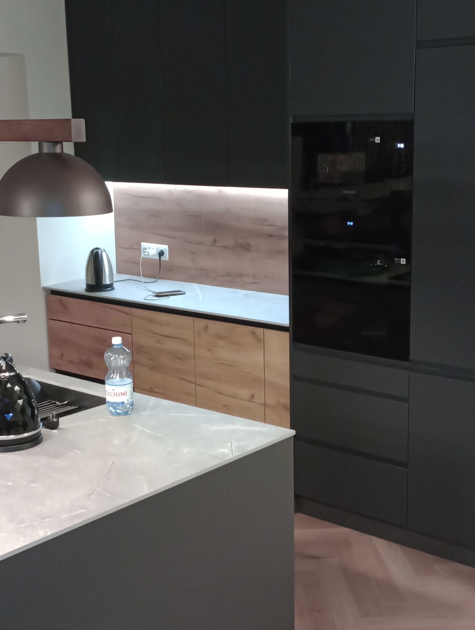 Nowoczesna kuchnia: czarne szafki, drewniane akcenty, marmurowy blat wyspy. Oświetlenie LED podkreśla unikalny design. Widoczny czajnik i butelka wody.