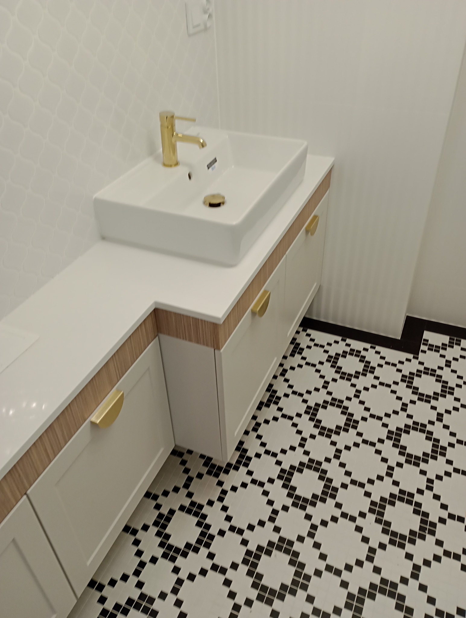 Biała szafka łazienkowa na wymiar z blatem, złotą armaturą i uchwytami, na tle wzorzystej podłogi. Umywalka prostokątna, nablatowa.