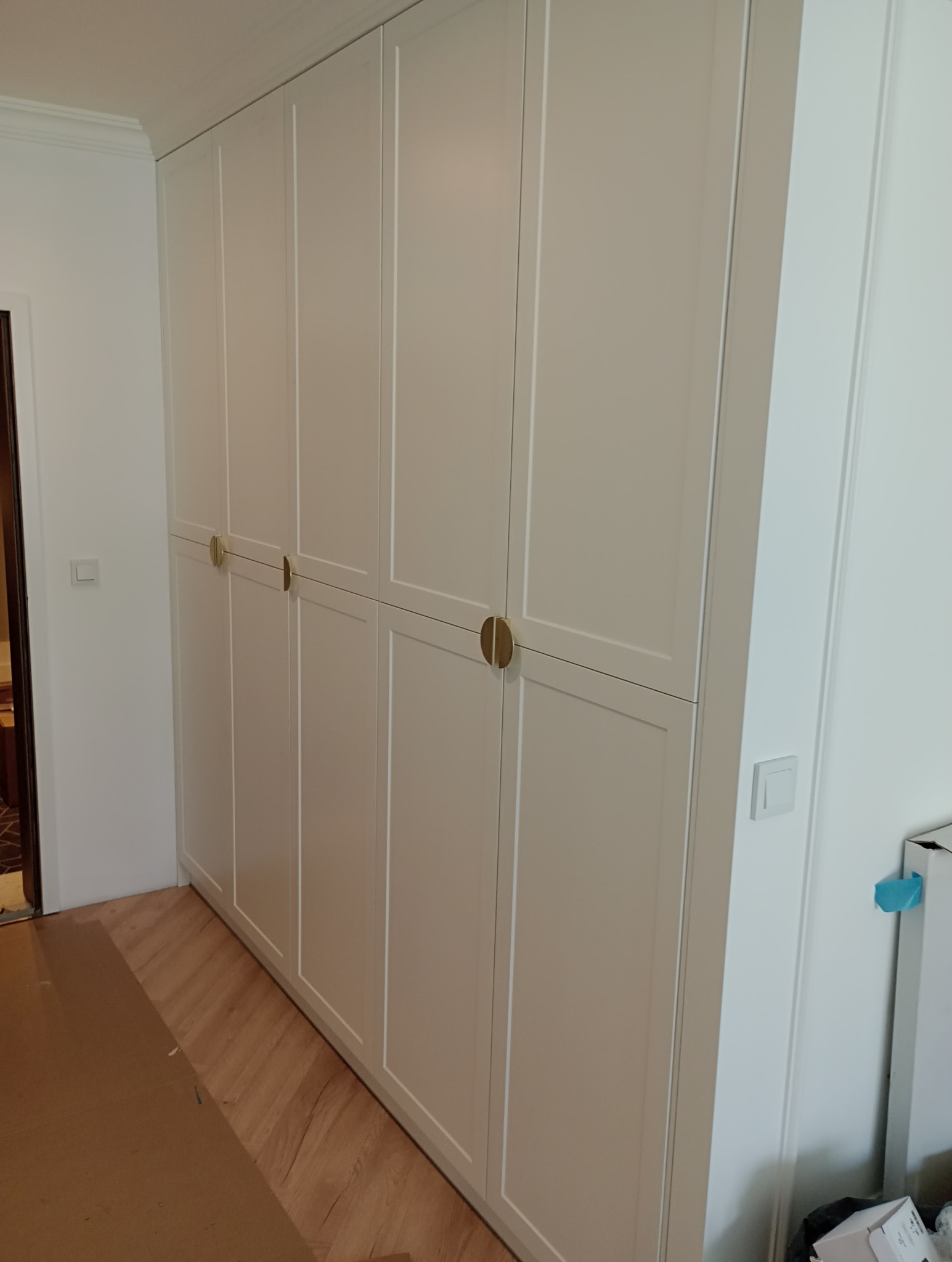 Elegancka, biała szafa wnękowa z subtelnymi, złotymi uchwytami, dopasowana do wnętrza z parkietem. Widoczne detale wykończenia i minimalistyczny design.