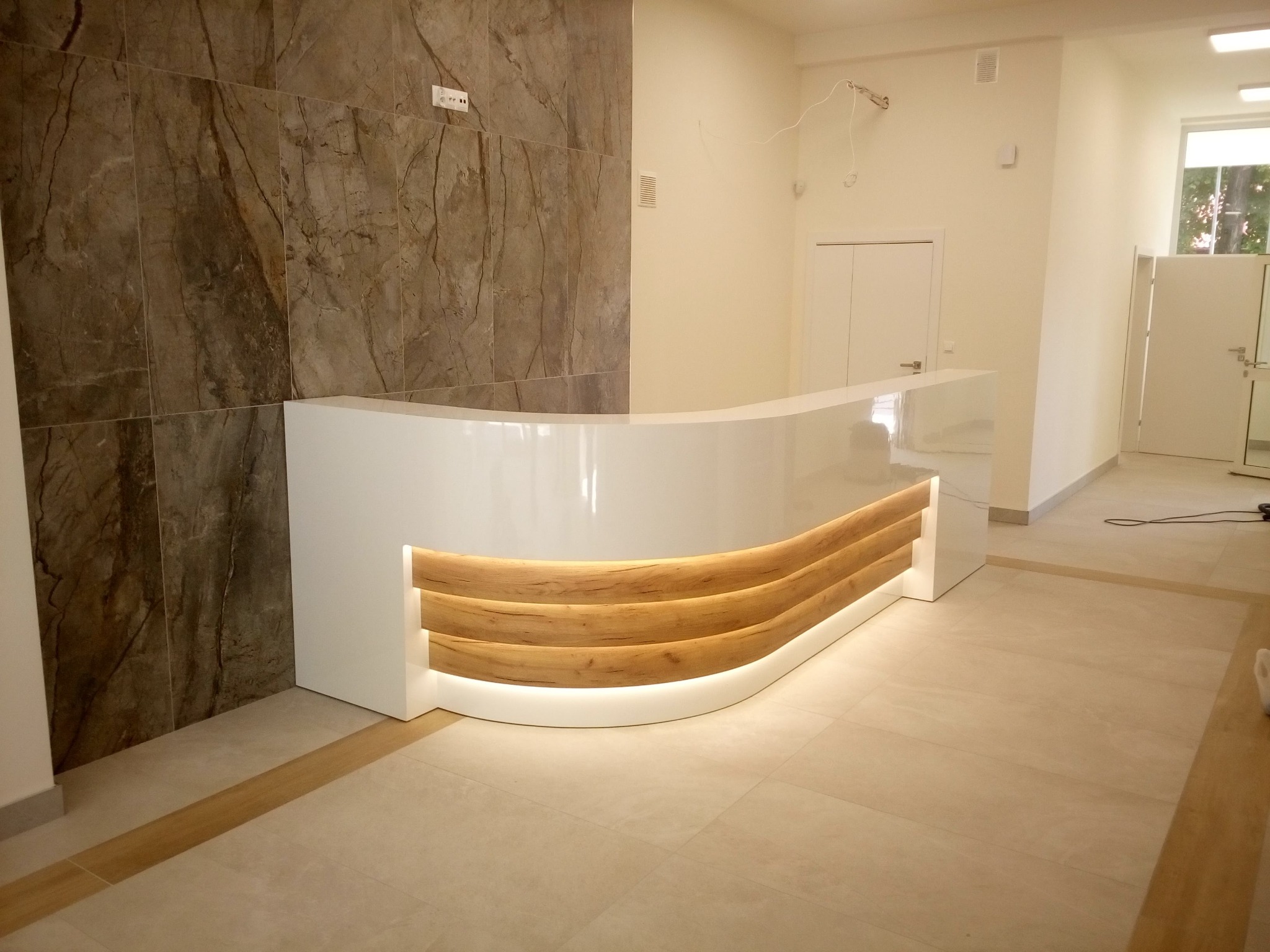 Błyszcząca, biała lada recepcyjna z elementami drewna i podświetleniem LED na tle marmurowej ściany. Widok na jasne wnętrze z drzwiami.