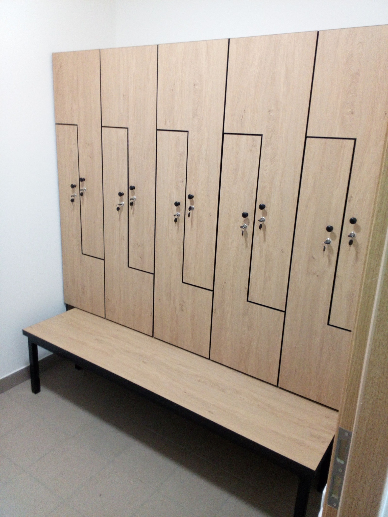 Jasne szafki z drewna z czarnymi detalami i ławką w szatni. Szafki zamykane na klucz, widoczne zamki z kluczykami. Prosty design, funkcjonalne rozwiązanie do przechowywania.