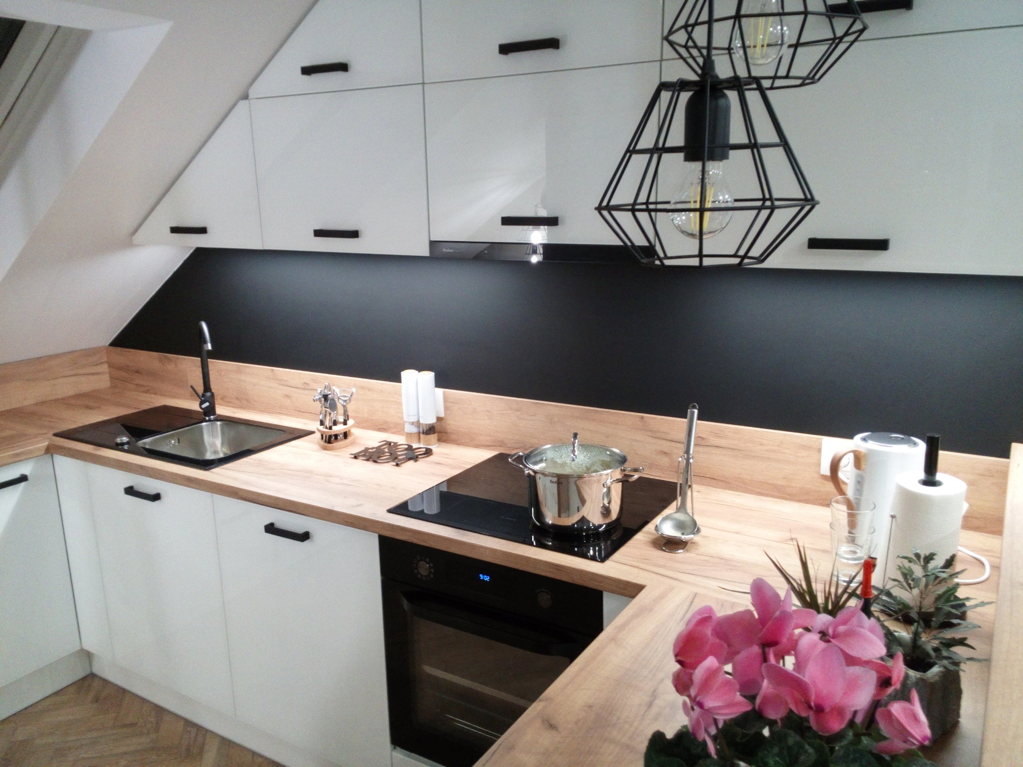 Nowoczesna kuchnia z białymi szafkami, drewnianym blatem i czarnym panelem. Nad blatem wiszą designerskie lampy. Na blacie kwiaty i akcesoria kuchenne.