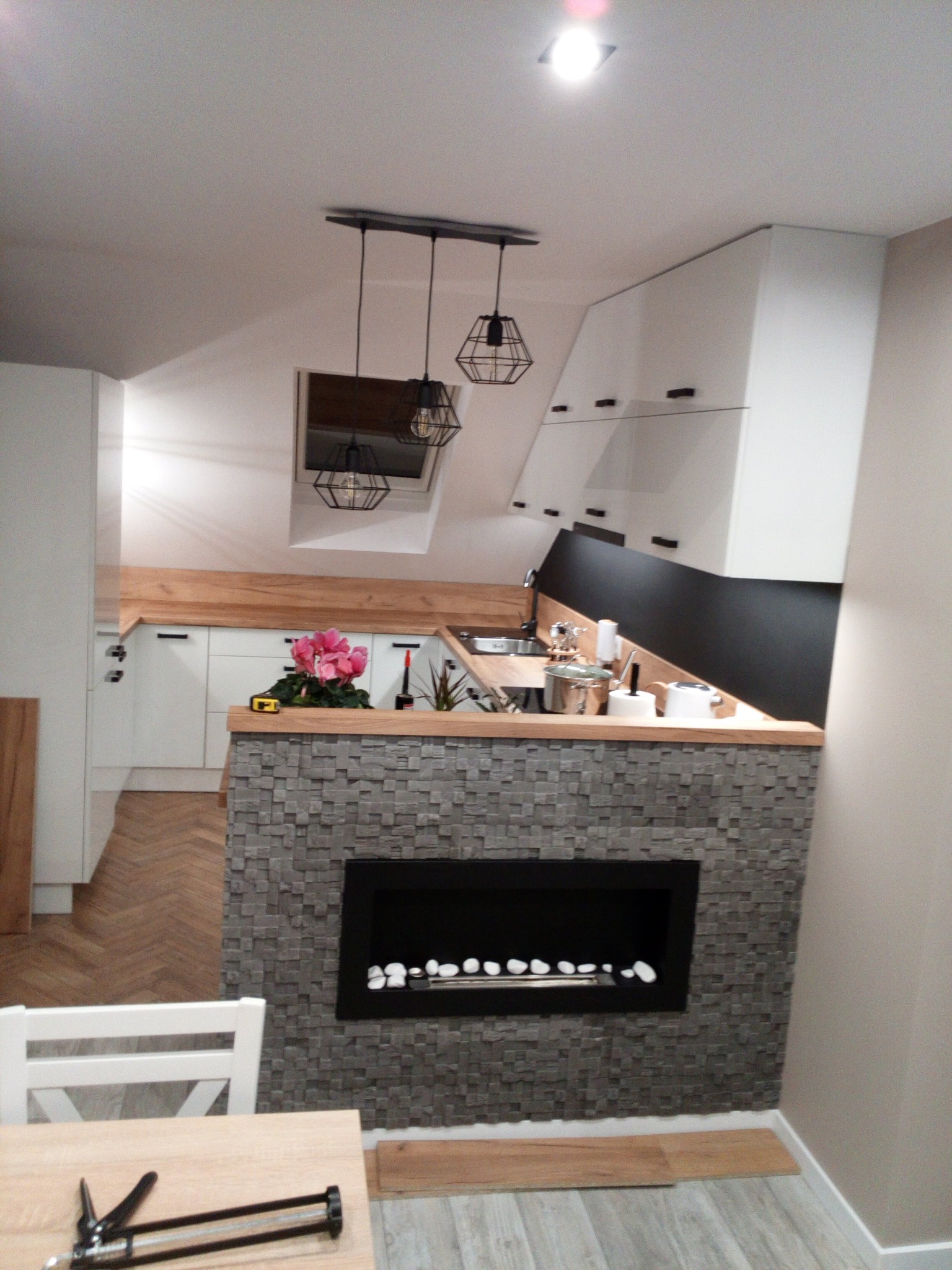 Nowoczesna kuchnia z wyspą wykończoną mozaiką i kominkiem elektrycznym, oświetlona wiszącymi lampami. Ukończona zabudowa kuchenna w skosach poddasza.