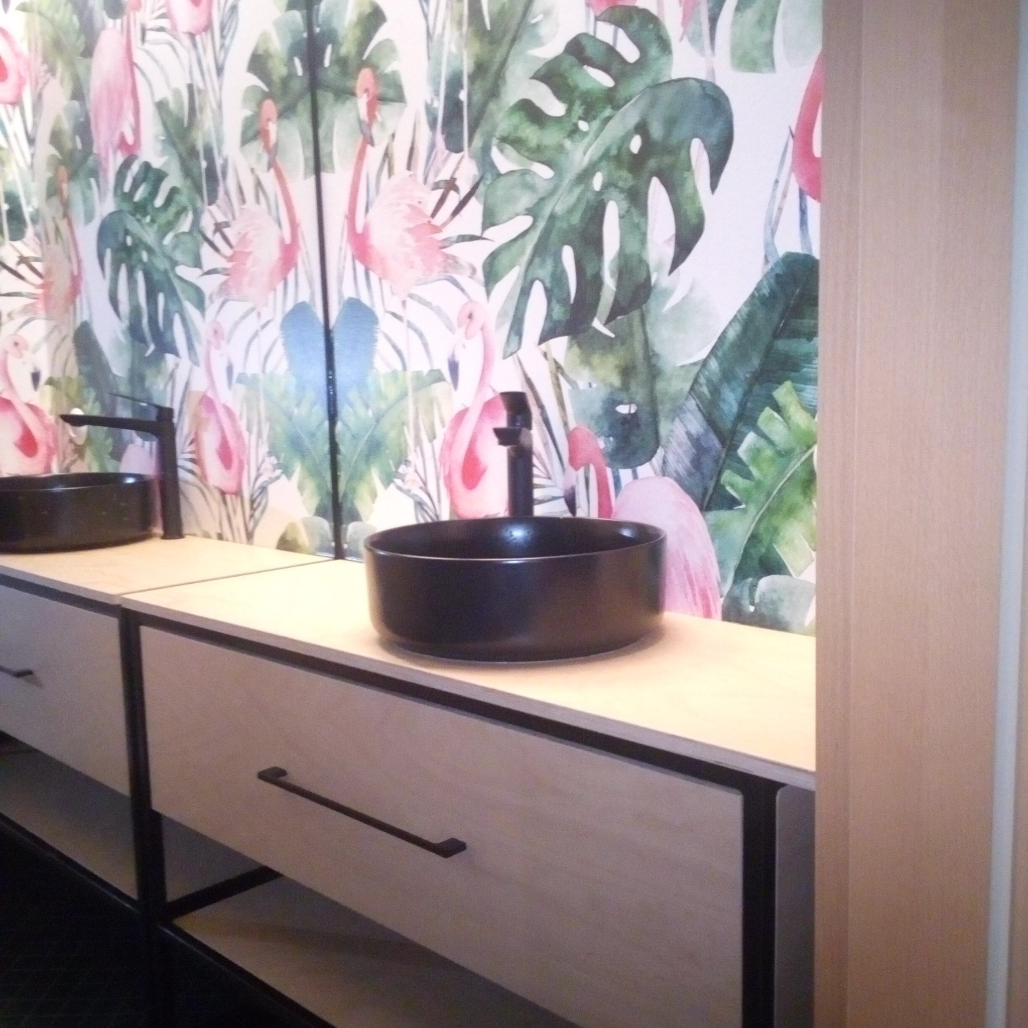 Nowoczesna szafka łazienkowa z czarną umywalką na blacie, na tle ściany z motywem flamingów i tropikalnych liści. Minimalistyczny design.