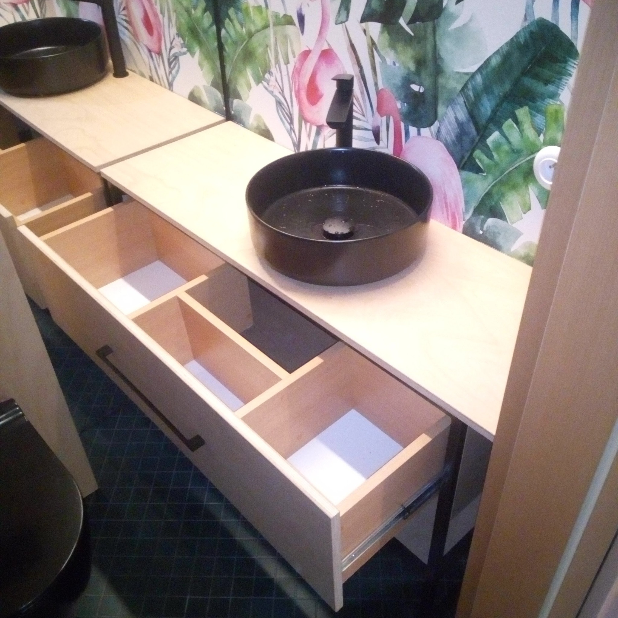 Biała, drewniana szafka łazienkowa z czarnymi umywalkami na blacie i otwartymi szufladami, na tle tapety w flamingi i liście.