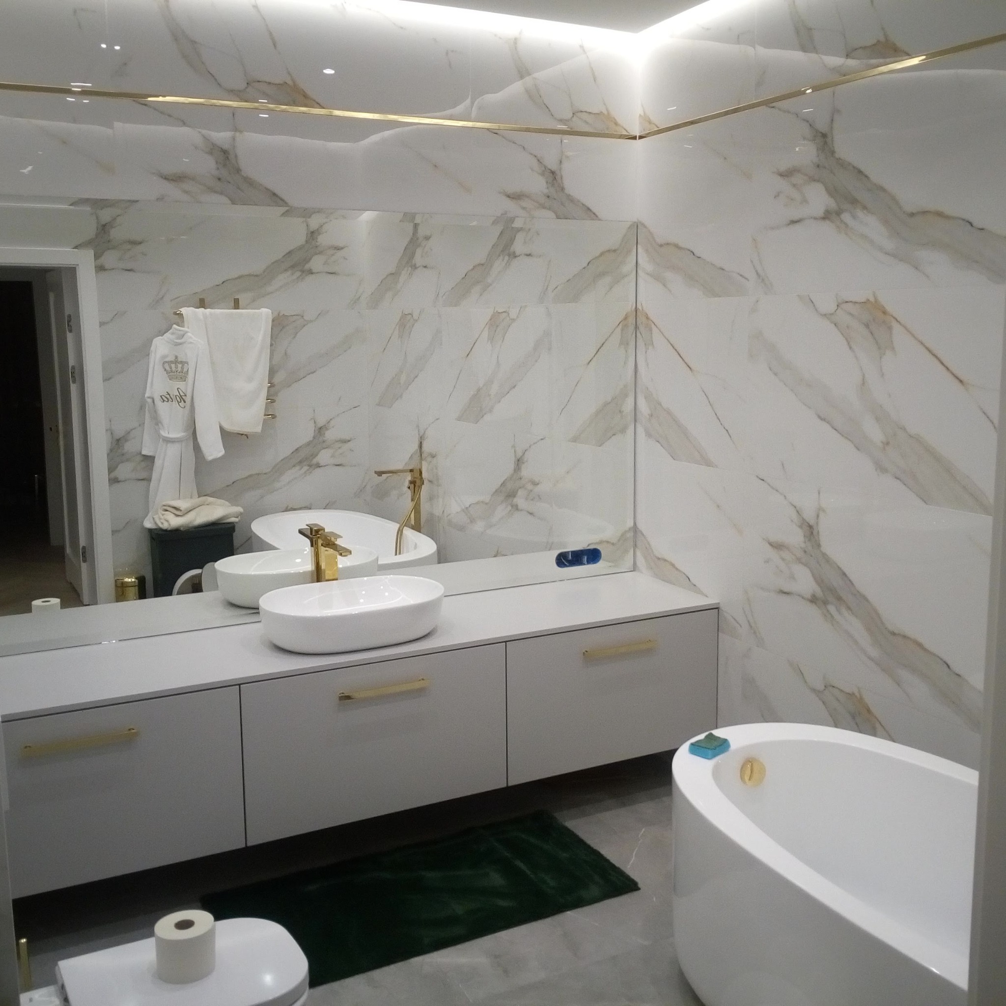 Elegancka łazienka z marmurowymi płytkami, białą szafką pod umywalkę z złotymi uchwytami i owalną wanną. Widoczne lustro i akcesoria łazienkowe.