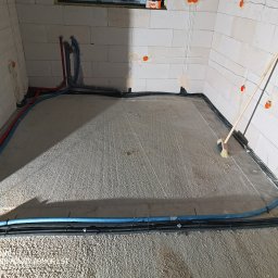 ETP Solution - Wylewka betonowa z rozprowadzonymi rurami ogrzewania podłogowego i kablami elektrycznymi w surowym stanie, przygotowywana do dalszych prac wykończeniowych, z widoczną szczotką do zamiatania.